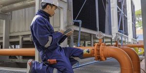 A technician in a hard hat uses a digital tablet during a commercial backflow inspection near large industrial plumbing pipes.
