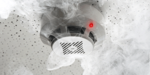 A white residential smoke detector mounted on a textured ceiling, active with a glowing red light and completely surrounded by thick white smoke curls.